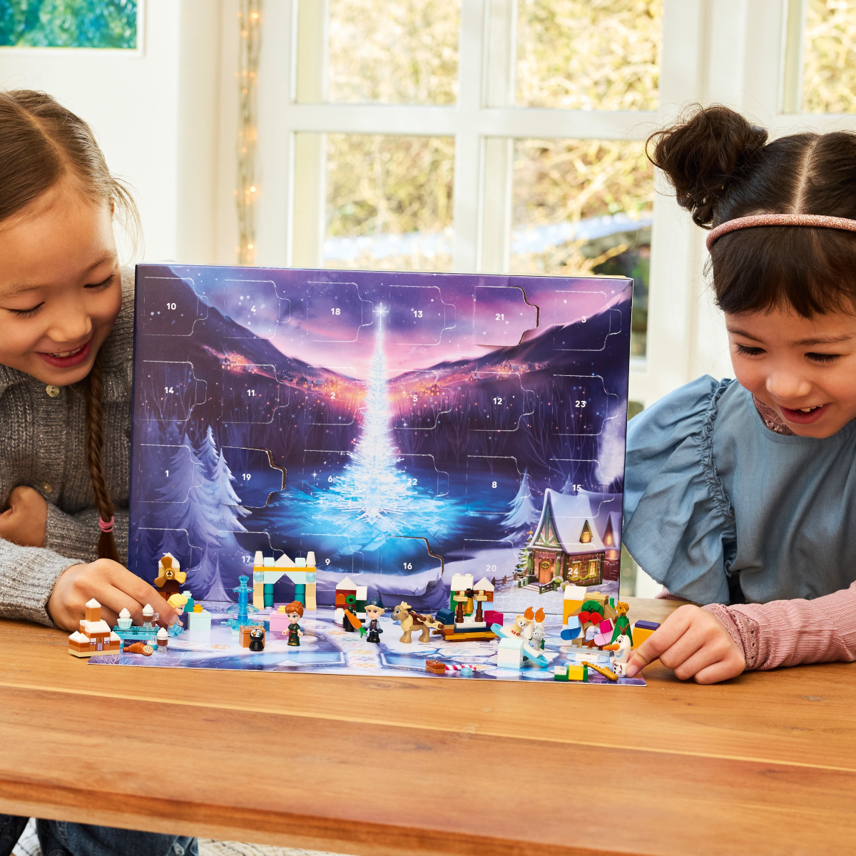 Two children, holding the LEGO Disney Frozen advent calendar while playing with the figures from it, on a wooden table.
