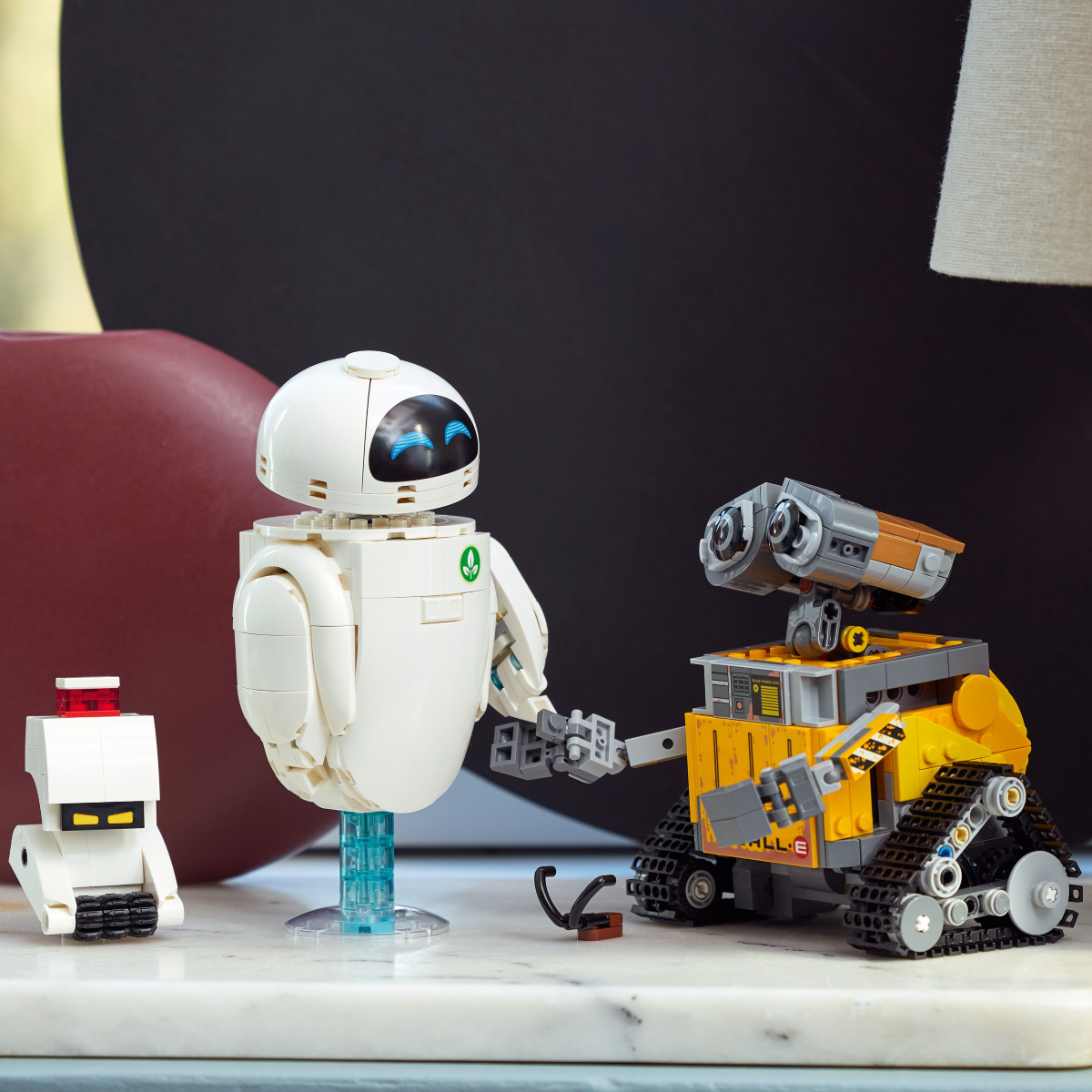 Two robot LEGO toys, one resembling WALL-E and the other resembling EVE from 'EVE,'holding hands on a marble table.